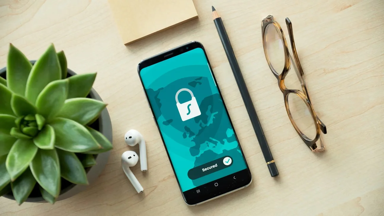 A smartphone displaying a turquoise screen with a white padlock icon rests on a light wooden desk, accompanied by a potted succulent, wireless earbuds, and a pencil.